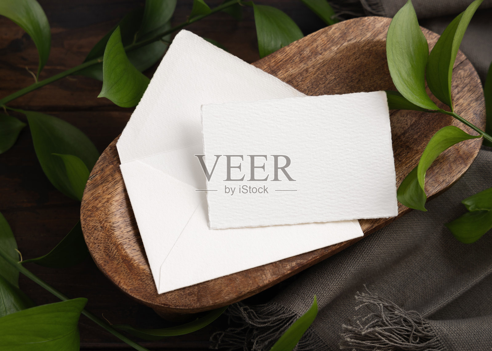 俯视视角，卡片和信封模型放在棕色木托盘上，旁边有绿叶和灰色织物。照片摄影图片
