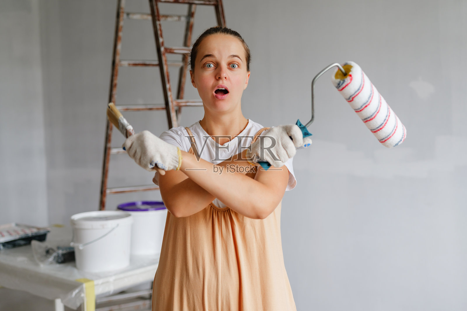一位女士拿着油漆用品，在准备粉刷房间墙壁时显得很惊讶。房间内有梯子和油漆桶在附近。照片摄影图片