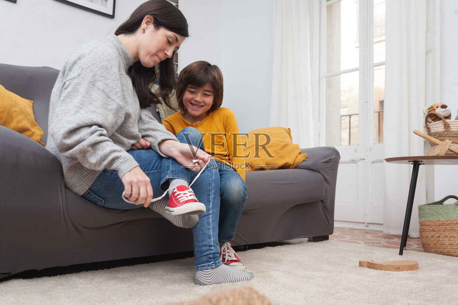 母亲在客厅沙发上帮助儿子脱掉红色运动鞋照片摄影图片