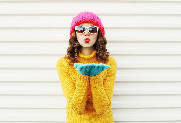 肖像时尚的女人吹红色的嘴唇，使送飞吻戴彩色手套针织帽在白色的背景图片下载