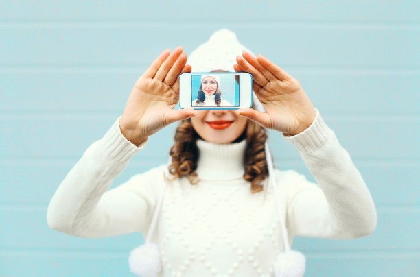 快乐的漂亮女孩拍照自拍照在蓝色背景的智能手机在冬季针织衣服图片下载