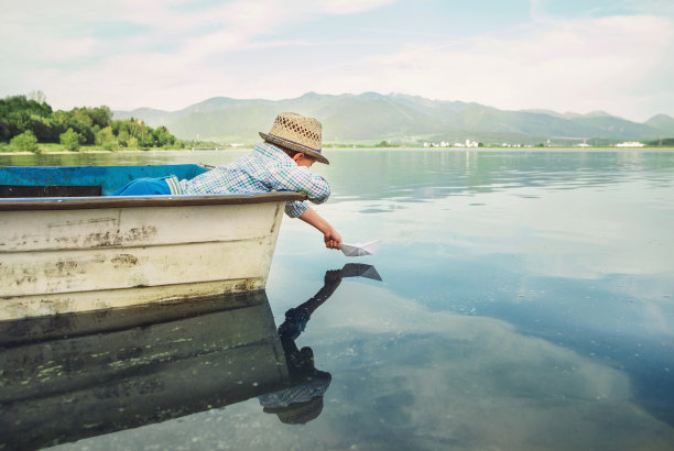 男孩从湖上的一条旧船上放纸船下水图片下载