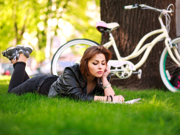 年轻女子骑着自行车在公园的草地上看书图片下载