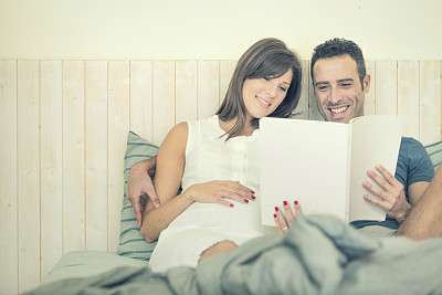 一对幸福的怀孕夫妇在床上看书图片下载