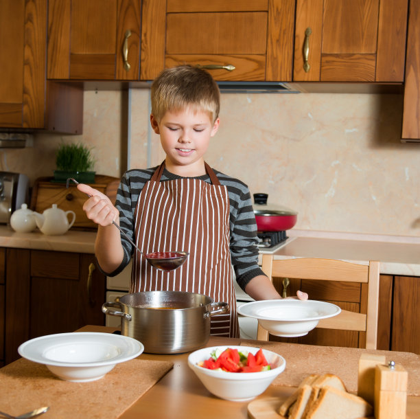 快乐的男孩在厨房里把汤倒进盘子里。图片下载