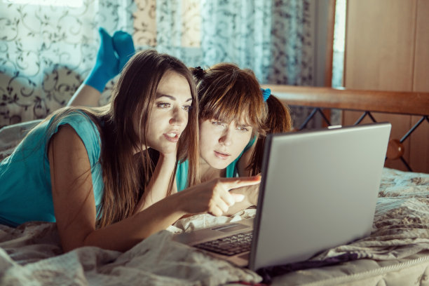 两个穿着蓝色t恤的女孩在床上用笔记本电脑。图片下载