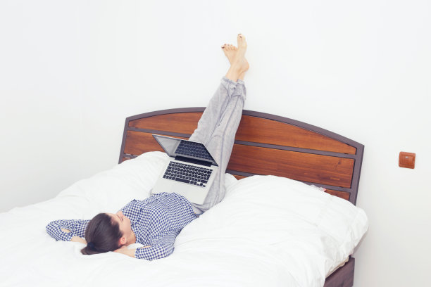 舒适的年轻女人坐在床上拿着笔记本电脑。图片下载