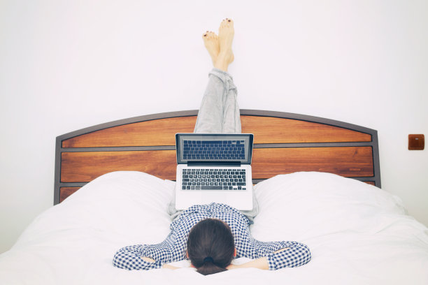 舒适的年轻女人坐在床上拿着笔记本电脑。图片下载