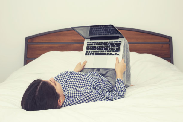 舒适的年轻女人坐在床上拿着笔记本电脑。图片下载