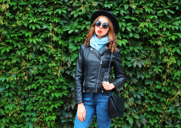 时尚漂亮的年轻女子穿着黑岩夹克，帽子和包在绿色树叶纹理墙图片下载