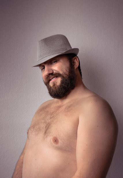 英俊的半裸胡子男人，戴着灰色的帽子，站在灰色的背景墙上。图片下载