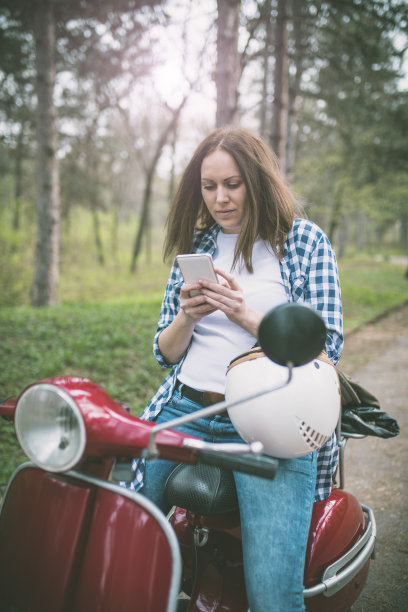 年轻的潮女人戴着头盔坐在老式摩托车上，用手机自拍图片下载