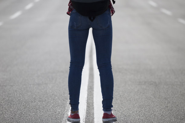女人的腿上穿着牛仔裤，就在道路的分界线上。后面的观点。图片下载