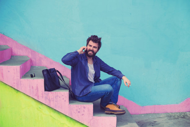 留着胡子的帅气嬉皮士一边打电话一边坐在楼梯上图片下载