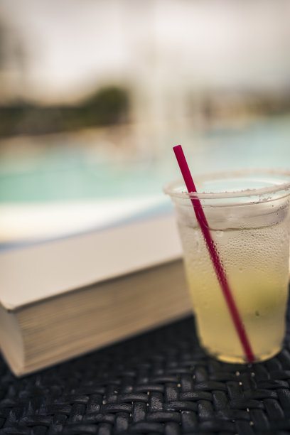 这张照片是在一个游泳池旁拍摄的，镜头里的margarita和红色吸管边缘上的盐和凝结在一次性杯子上清晰可见。桌子上也有一本书。图片下载