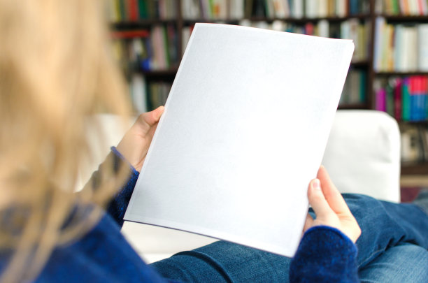 一个金发女人坐在家里的客厅里，手里拿着一本空白的小册子，上面还有复印的地方。素材图片