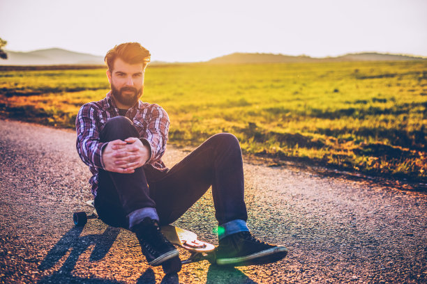 在一个阳光明媚的夏日里，坐在滑板上的帅气大胡子嬉皮士图片下载
