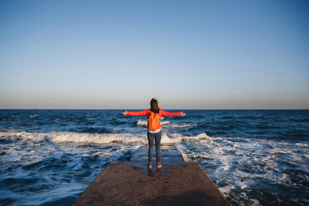 年轻快乐的女孩背着背包站在海边呼吸新鲜空气，举起双臂欣赏风景图片下载