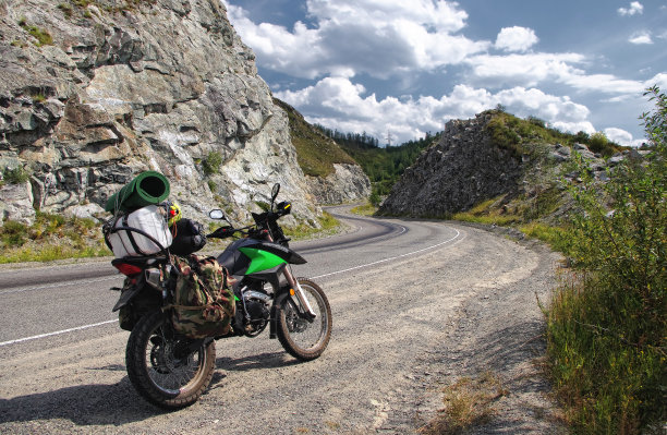 摩托车旅行家带着行李箱站在柏油路上，高原上有绿色的草地，在蓝天下的阿尔泰山脉俄罗斯西伯利亚图片下载