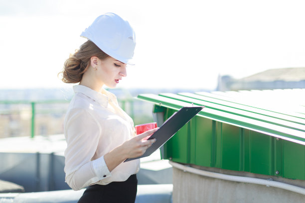 漂亮的女商人穿着白衬衫、手表、头盔和黑裙子站在屋顶上，拿着平板和一杯咖啡图片下载