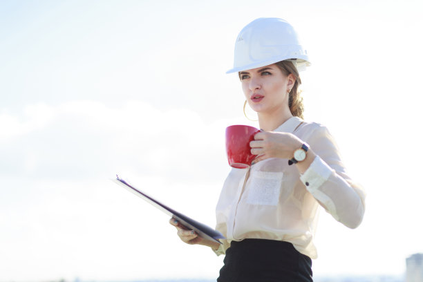 漂亮的女商人穿着白衬衫、手表、头盔和黑裙子站在屋顶上，拿着平板和一杯咖啡图片下载