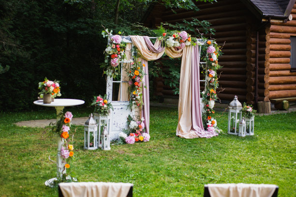 美丽的户外婚礼。装饰好的椅子放在草地上。婚礼拱门由布料和白色和粉红色的花朵在绿色的自然背景。旧门，乡村风格。图片下载