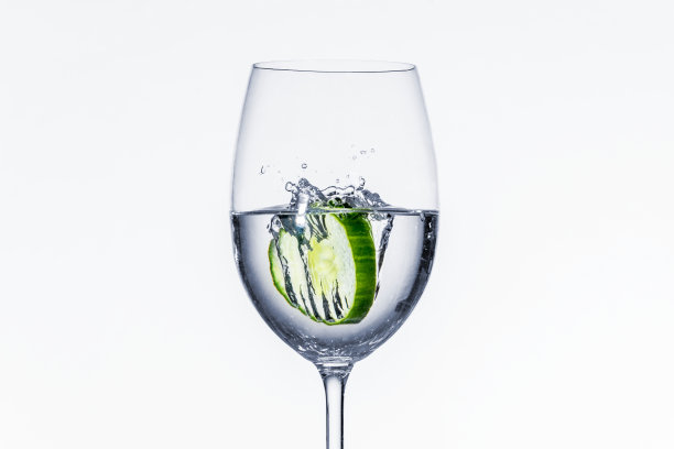 在白色背景下，黄瓜片落入玻璃杯中图片下载