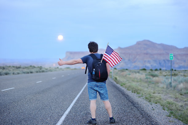 背包里装着美国国旗的中年男性游客沿着一条荒凉的道路搭便车图片下载