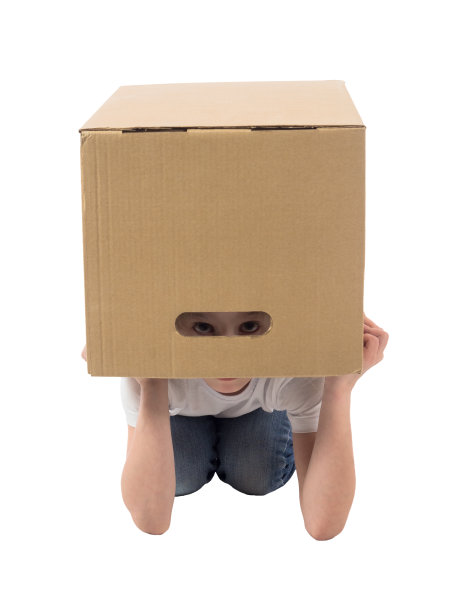 穿着白色t恤和牛仔裤的小女孩躲在一个盒子里，透过白色背景上的插槽向外偷看图片下载