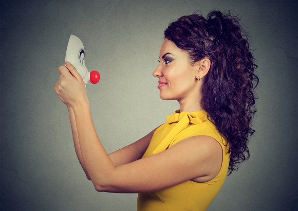 微笑的女人看着小丑面具孤立在灰色的墙壁背景图片下载