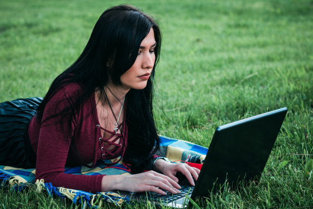 年轻美丽的女人躺在公园的草地上使用笔记本电脑。图片下载