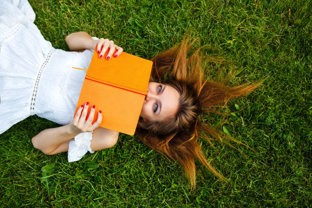 女孩把脸藏在草地上的书后面。图片下载