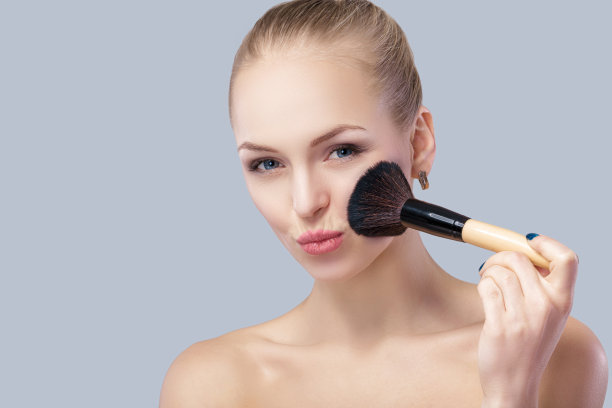 美丽的金发女人拿着化妆刷在灰色的背景。图片下载