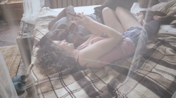 年轻的多种族夫妇穿着睡衣躺在床上看照片。男人和女人都在使用智能手机。视图通过窗帘图片下载