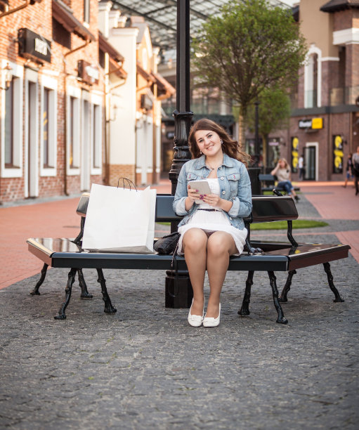 女人坐在长凳上，拿着购物袋，用着平板电脑图片下载