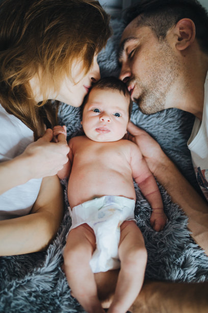 幸福的家庭。年轻的父母躺在床上亲吻可爱的新生婴儿。图片下载