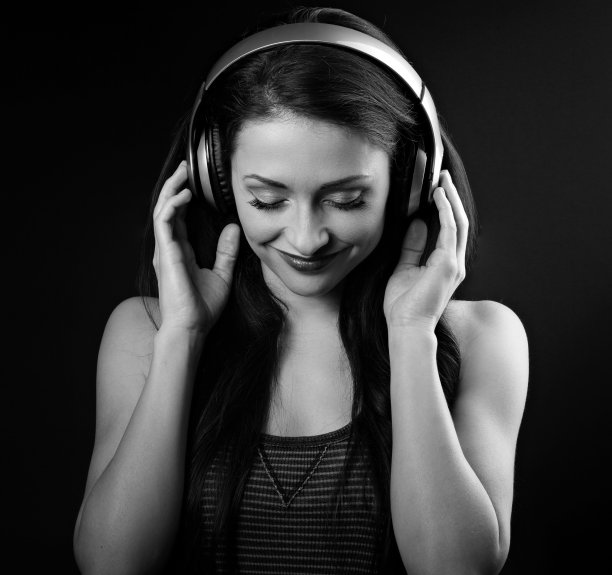 美丽的享受年轻女子听音乐在无线耳机与闭上眼睛在黑暗的阴影背景。特写黑白肖像图片下载