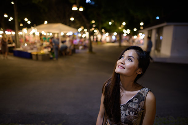 年轻快乐美丽的亚洲女人微笑和思考在泰国华欣夜市与人们买卖各种商品图片下载