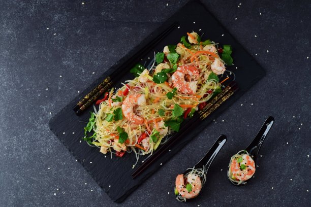 在灰色抽象背景上，用传统汤勺盛着黑色石盘上的虾仁和蔬菜面条。健康食品的概念图片下载