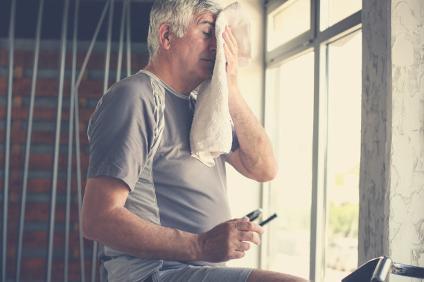 老人坐在健身机上。男子用毛巾擦脸。锻炼在健身房。素材图片