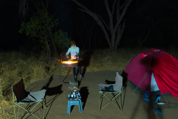 妇女晚上用柴火和烤肉设备做饭。前景是帐篷和椅子。在非洲国家公园探险。图片下载