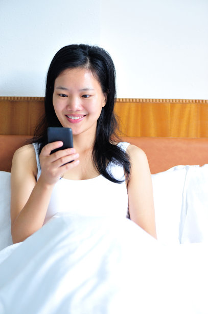 微笑的女人靠在床上用她的智能手机图片下载