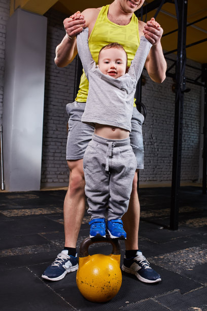 快乐的父亲牵着儿子的手，儿子站在体育馆砖墙上的壶铃上。图片下载
