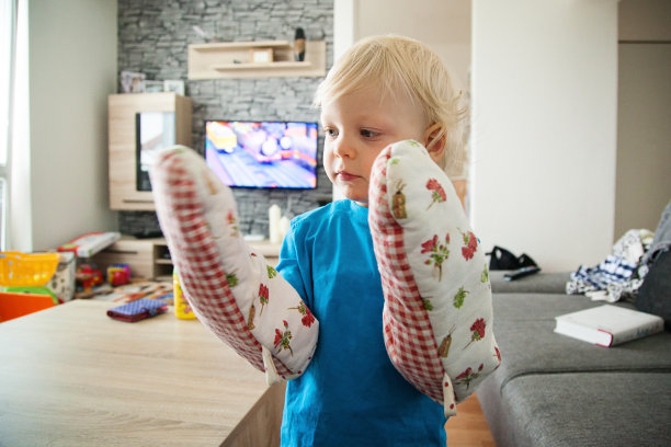可爱的金发小男孩站在家里，戴着成人尺寸的烤箱手套，看着自己的手图片下载