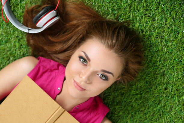 年轻美丽的女孩躺在公园的草地上，手里拿着书图片下载