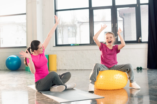 快乐的妈妈坐在瑜伽垫上，女儿在健身球上在健身房玩图片下载