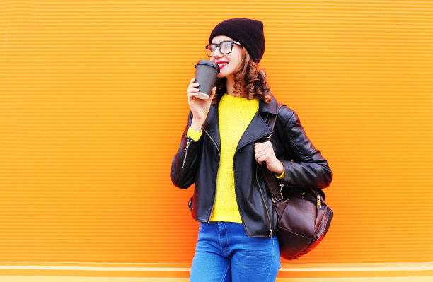 时尚年轻漂亮的年轻女人喝咖啡从杯戴黑帽子，摇滚夹克在五颜六色的橙色背景图片下载