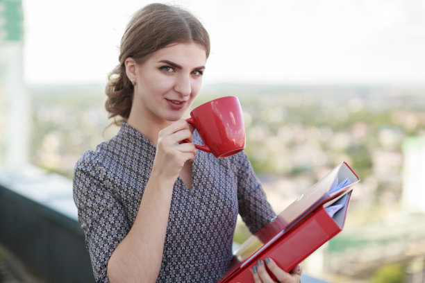 漂亮的女商人，穿着花式服装站在屋顶上，拿着文件夹和红杯子图片下载