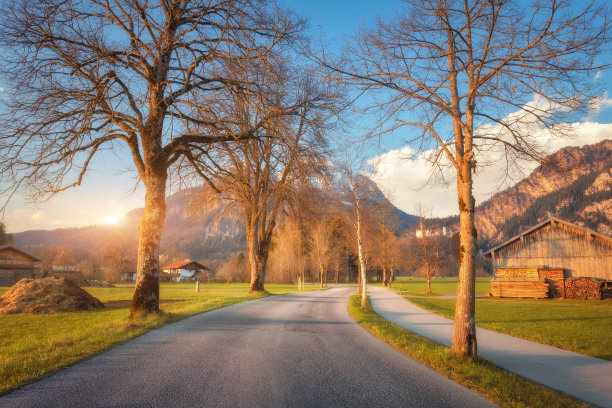 在阳光明媚的傍晚，绿树成荫的乡间小路、五彩缤纷的绿草、群山和蓝天白云，德国的春天。日落时乡间的柏油路。乡村的观点。旅游的背景图片下载