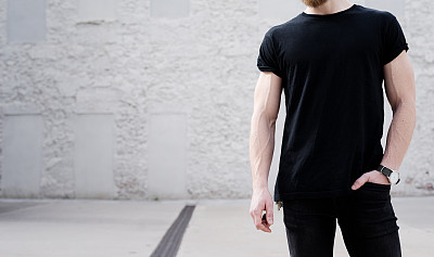 年轻的蓄着胡须的男子穿着黑色t恤和牛仔裤在外面摆姿势。背景是白色的砖墙。Hotizontal模型，正面视图图片下载